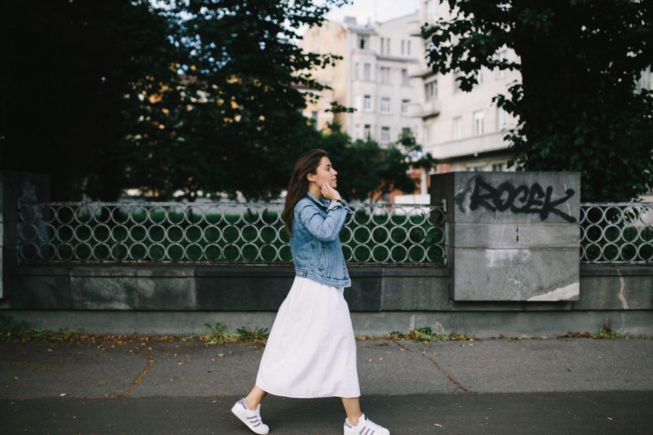 Full body side view of confident female in stylish outfit strolling on town street on walkway near stone barrier and tree with green foliage near buildings in daytime